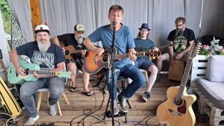 A group of men are playing guitars in a room. One man is sitting on a stool and singing into a microphone.