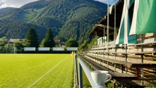 A serene view of a soccer field with empty bleachers, a cup of coffee on a railing, and a yellow flag in the background. The field is surrounded by lush greenery and mountains.