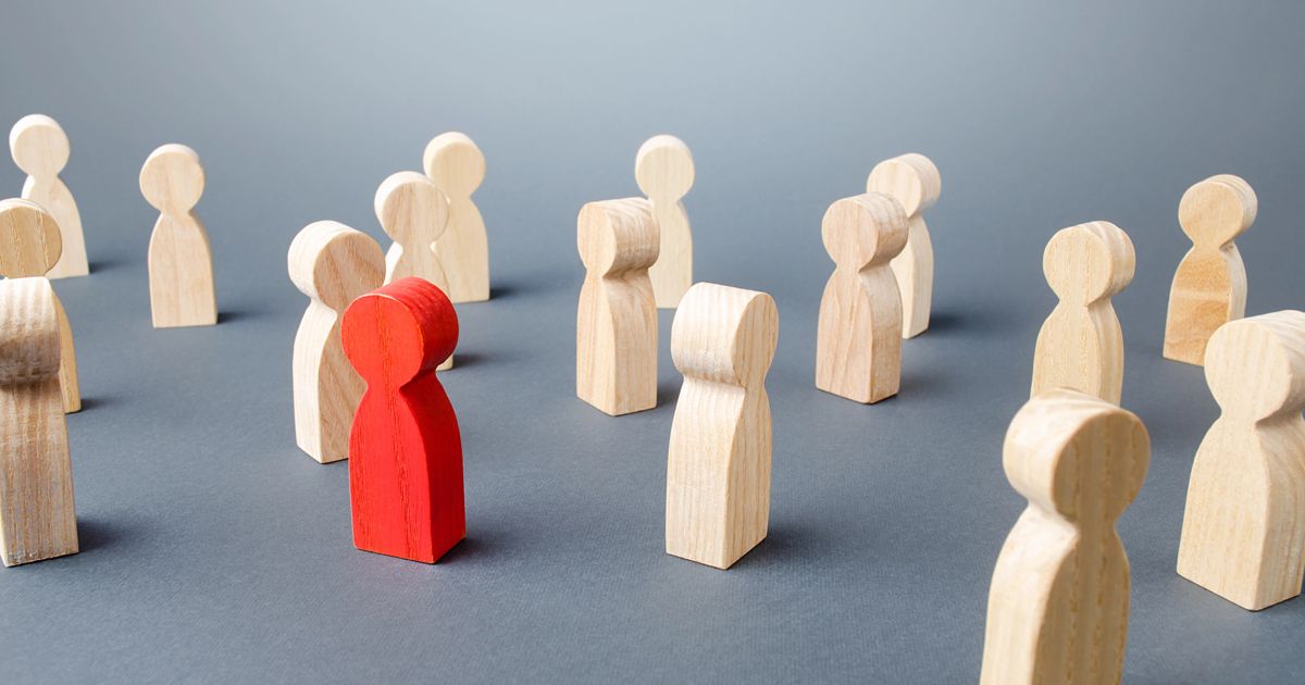 A red wooden figure stands out among a group of white wooden figures on a gray background.