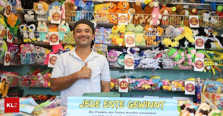 Ein Mann in einem weißen Hemd steht vor einem Geschäft voller Plüschtiere und gibt einen Daumen hoch. Das Geschäft hat ein Schild mit der Aufschrift 'Jeder Ent gewinnt' und Regale, die mit verschiedenen Spielzeugen bestückt sind.