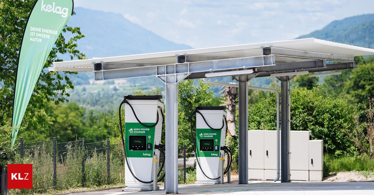 Two high-power electric vehicle chargers are installed under a metal canopy with solar panels on top. The chargers are green and white, with a digital display and a charging cable attached. The background features a scenic view of mountains and trees.