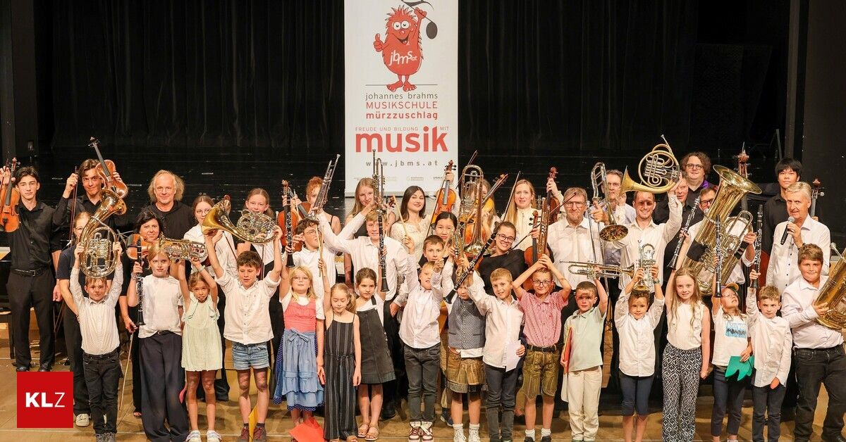 A group of children and adults on a stage, holding musical instruments, with a banner reading 'Johannes Brahms Musikschule Murzzuschlag' in the background.