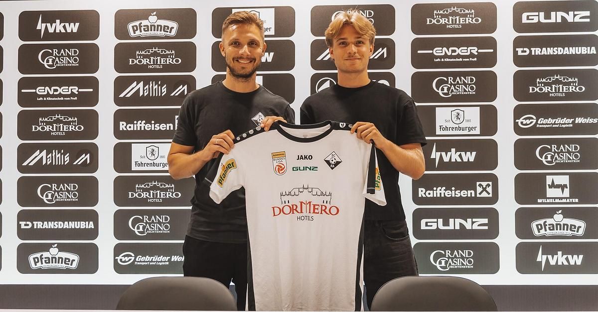Two men in black shirts stand in front of a wall with various logos, one holding a white soccer jersey with the word 'DORTMERO' on it.