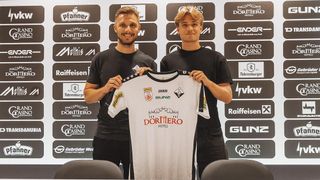Zwei Männer in schwarzen T-Shirts stehen vor einer Wand mit verschiedenen Logos, einer hält ein weißes Fußballtrikot mit dem Wort 'DORTMERO' darauf.