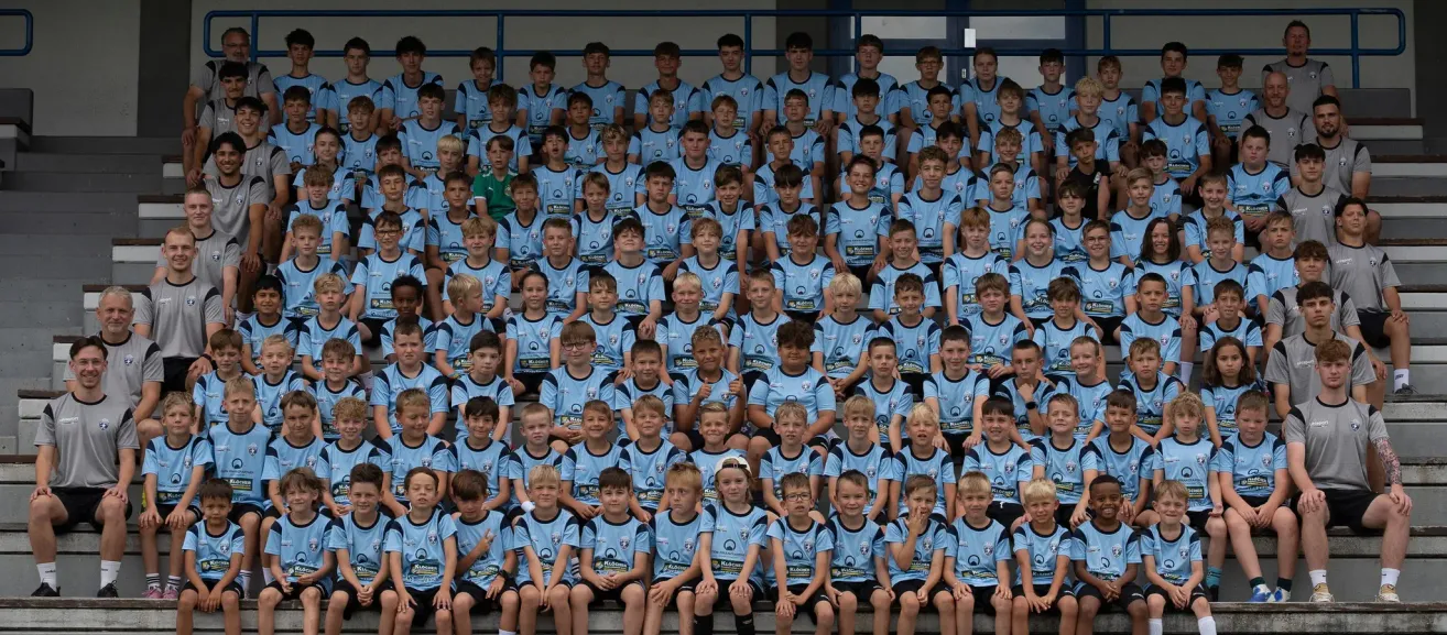 A group of young boys in blue jerseys with logos on the front, sitting on bleachers. They are smiling and posing for a photo.
