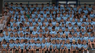 Eine Gruppe junger Jungen in blauen Trikots mit Logos auf der Vorderseite, die auf Tribünen sitzen. Sie lächeln und posieren für ein Foto.
