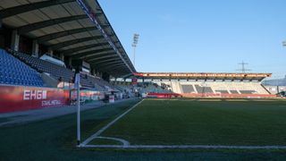 An empty stadium with a soccer field, empty bleachers, and a banner on the side. The sky is clear and blue.