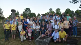 Eine große Gruppe von Menschen, lässig gekleidet, posiert für ein Gruppenfoto auf einer Wiese mit Bäumen im Hintergrund. Einige stehen, andere sitzen oder knien. Ein großer Stein ist im Vordergrund.