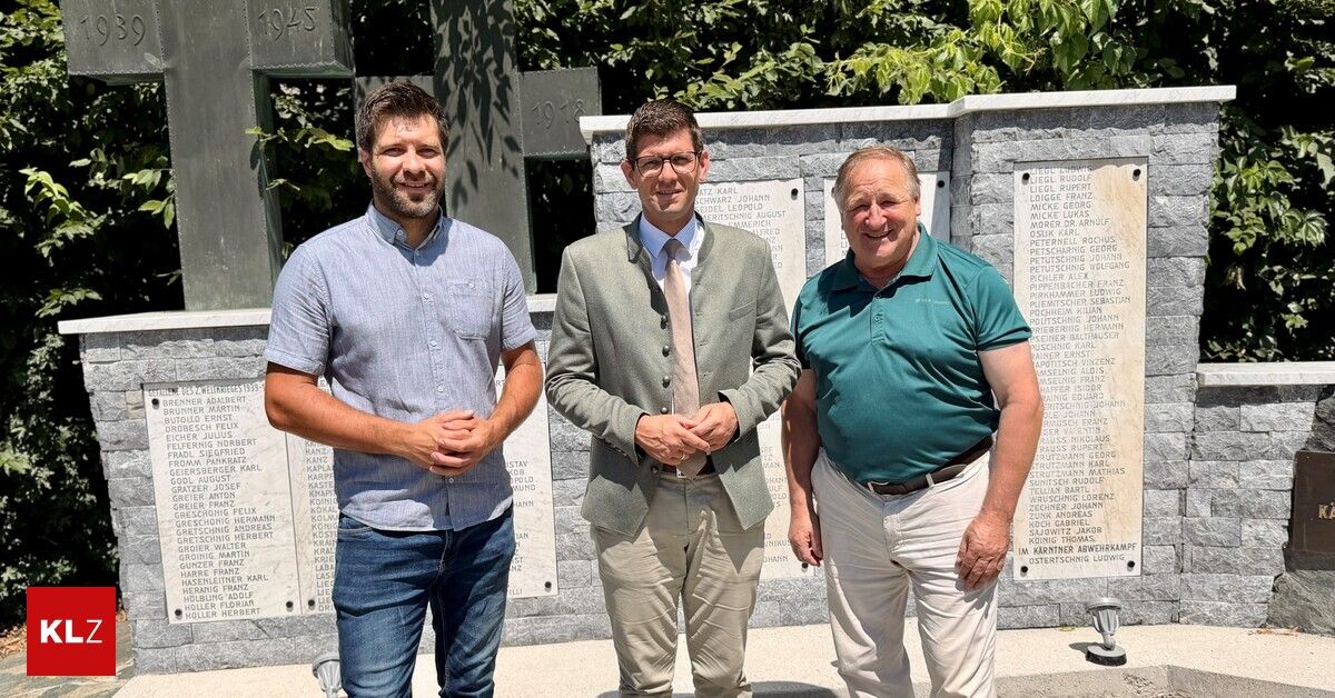 Three men stand in front of a memorial wall with engraved names and dates, smiling for a photo. The man on the left wears a blue shirt and jeans, the middle man a gray suit and tie, and the right man a green polo shirt.