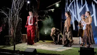 Drei Personen auf einer Bühne, einer in rotem Anzug, ein anderer kniet, und eine Frau in braunem Kleid. Der Hintergrund ist dunkel mit Bäumen und einem weißen Hintergrund.