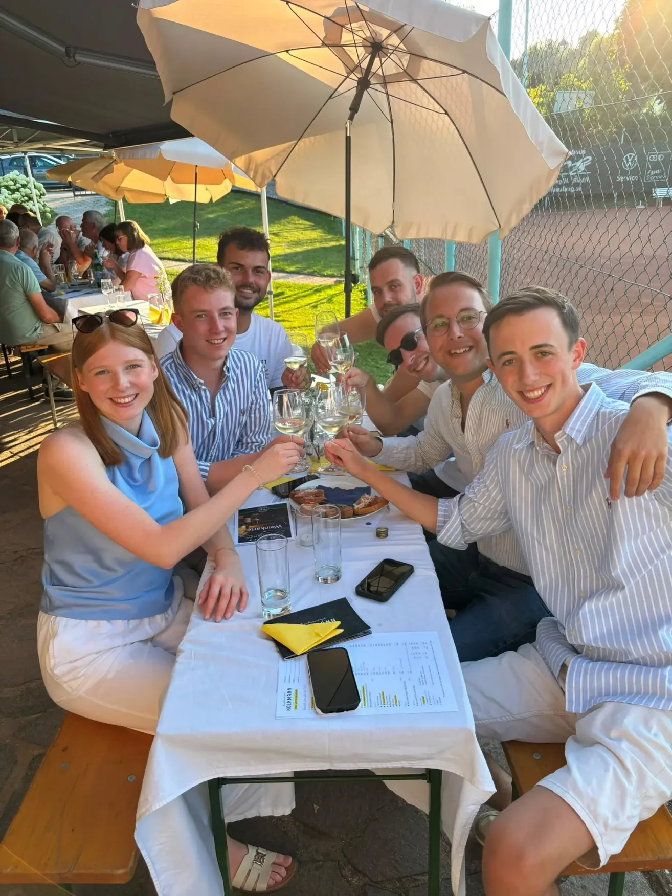 Eine Gruppe von Menschen, darunter Männer und Frauen, sitzt um einen Tisch in einer Außenumgebung und lächelt mit Weinläsern in der Hand. Sie scheinen ein Ereignis zu feiern.