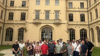 Eine Gruppe von Menschen in Freizeitkleidung steht vor einem großen, historischen Gebäude mit vielen Fenstern und einem großen Eingang.