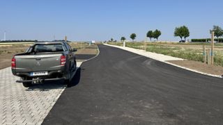 Ein Pickup-Truck ist am Rande einer frisch asphaltierten Straße geparkt, mit einem klaren blauen Himmel darüber und etwas Grün und Bäumen im Hintergrund.