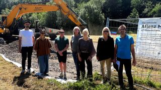 Eine Gruppe von Menschen in Freizeitkleidung steht vor einer Baustelle mit einem gelben Bagger im Hintergrund. Die Baustelle ist von einem Metallzaun umgeben, und der Boden ist mit Kies bedeckt.