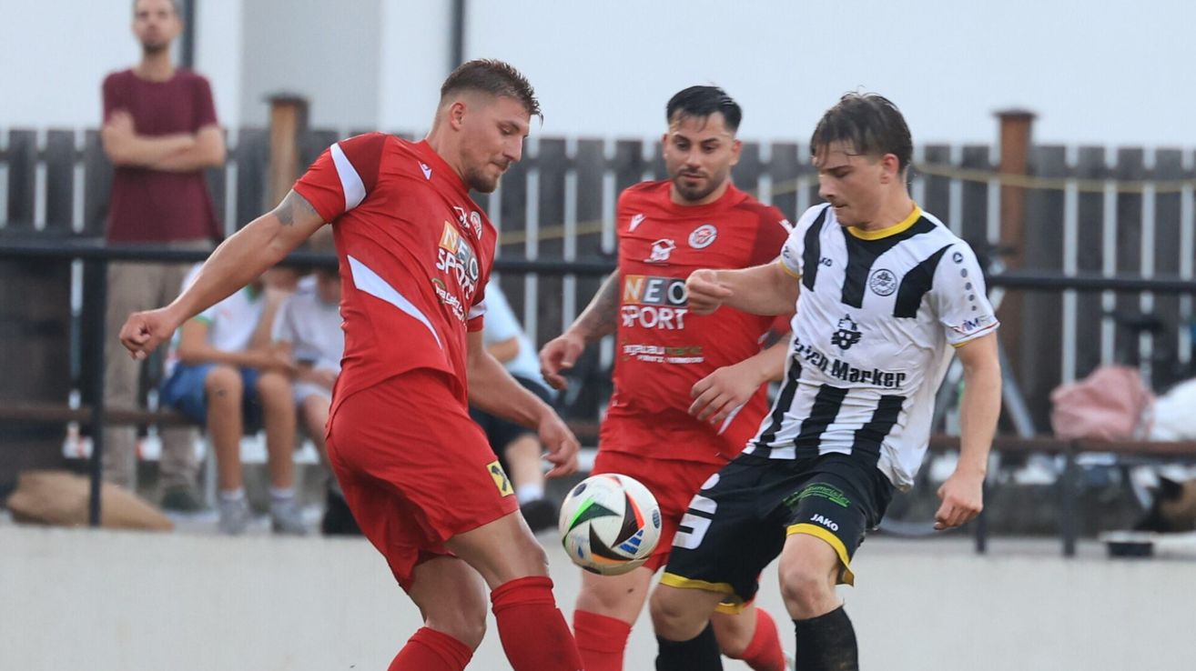 Drei Fußballspieler in roten, weißen und schwarzen Trikots sind in einem Spiel. Der Spieler in Rot schaut auf den Ball.