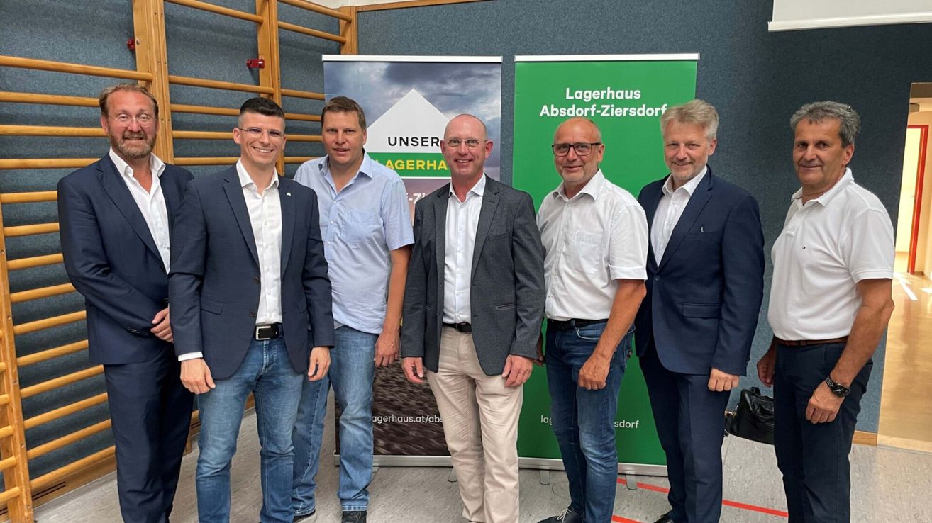 Fünf Männer in Geschäftskleidung stehen zusammen vor einem Banner mit dem Text 'Lagerhaus Absorf-Ziersdorf'. Der Mann in der Mitte trägt einen grauen Anzug und eine Brille.
