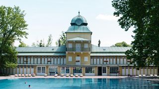 Ein gelbes Gebäude mit einem Uhrturm steht neben einem großen Schwimmbecken. Das Gebäude hat mehrere Fenster und Türen, und draußen stehen Bänke und Topfpflanzen. Eine Person steht am Pool.