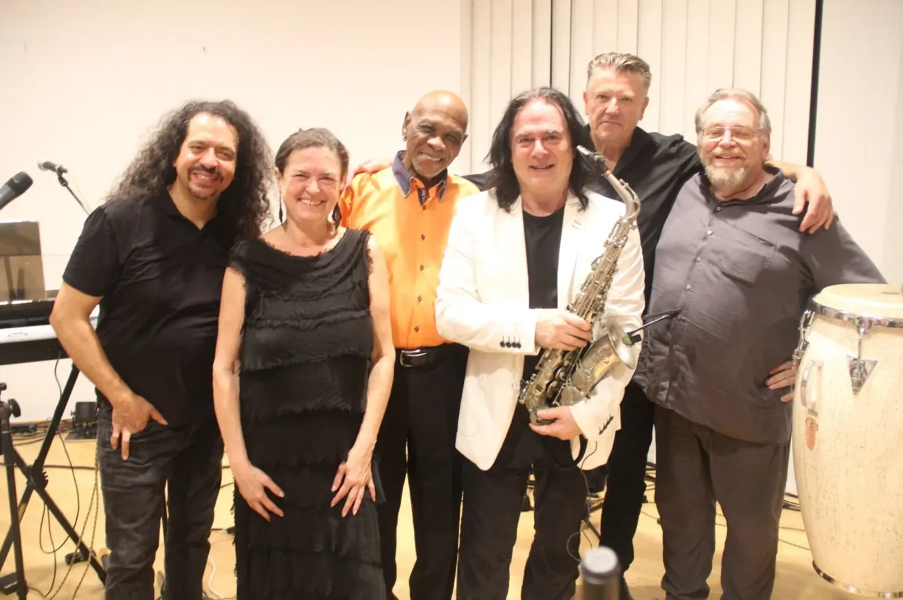 Eine Gruppe von Musikern, darunter eine Frau in einem schwarzen Kleid und ein Mann mit einem Saxophon, lächeln und posieren für ein Foto.