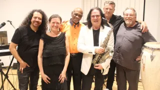 Eine Gruppe von Musikern, darunter eine Frau in einem schwarzen Kleid und ein Mann mit einem Saxophon, lächeln und posieren für ein Foto.