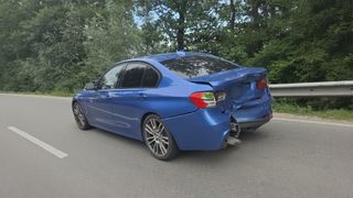 Ein blauer BMW mit beschädigtem Heck steht am Straßenrand. Im Hintergrund sind Bäume und Grünflächen zu sehen.