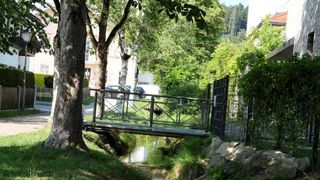 Eine kleine Brücke aus Metall und Holz überspannt einen schmalen Bach in einer üppigen, grünen Parkumgebung. Bäume und Pflanzen umgeben das Gebiet, ein Gebäude ist im Hintergrund sichtbar.