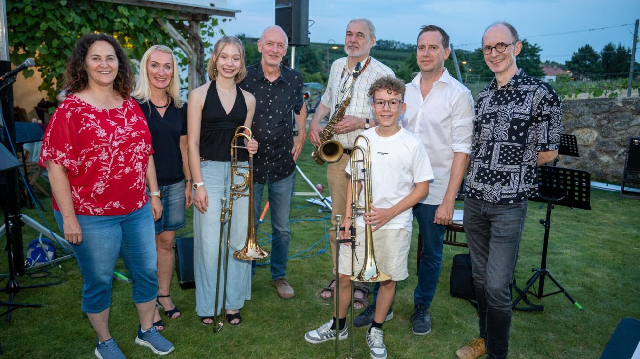 Eine Gruppe von Musikern und ihren Familien posiert für ein Foto auf einem Rasenfeld, mit Musikinstrumenten wie Posaunen und Saxophonen.