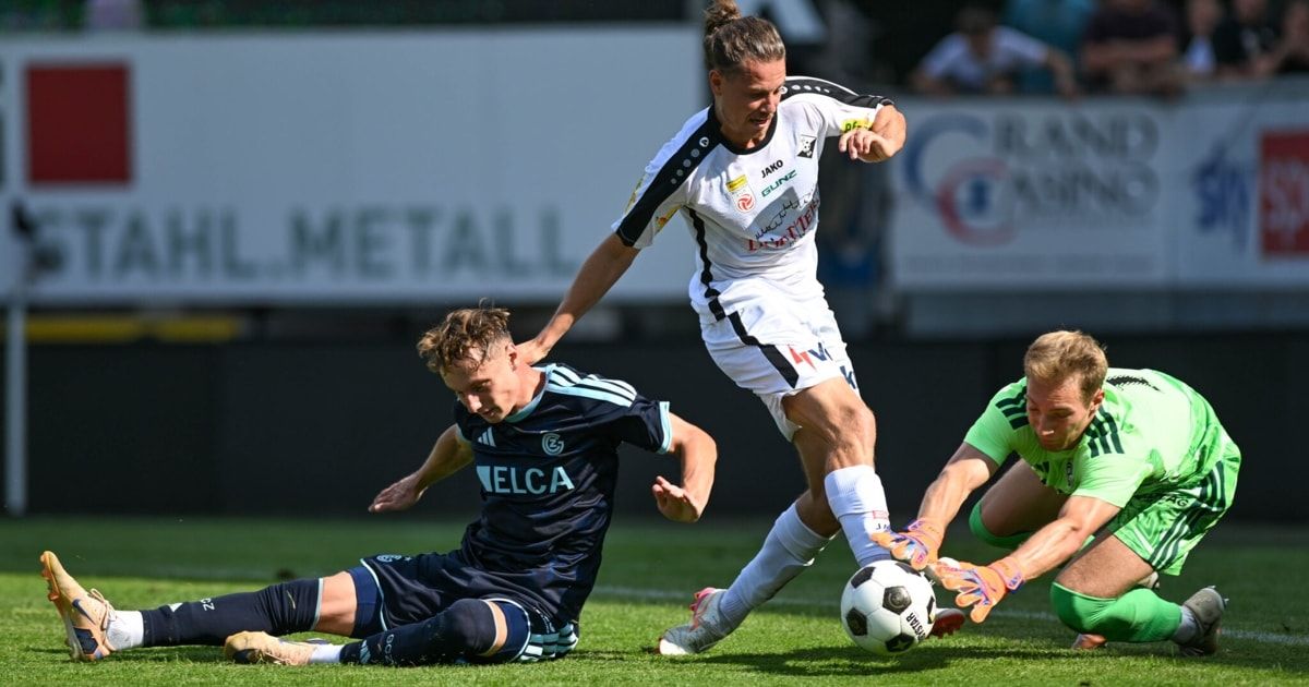 Drei Fußballspieler sind auf dem Spielfeld in Aktion. Ein Spieler in Weiß versucht, dem Spieler in Blau den Ball zu stehlen, während der Spieler in Grün versucht, das Spiel zu blockieren.