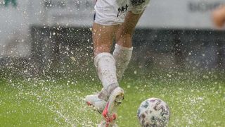 Ein Fußballspieler in weißen Shorts und Socken tritt einen Fußball, wodurch Wasserspritzer entstehen.