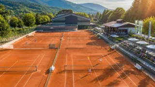 Ein Tennisplatz mit zwei Spielern in Aktion, umgeben von einem Gebäude und Bäumen, mit Bergen im Hintergrund.