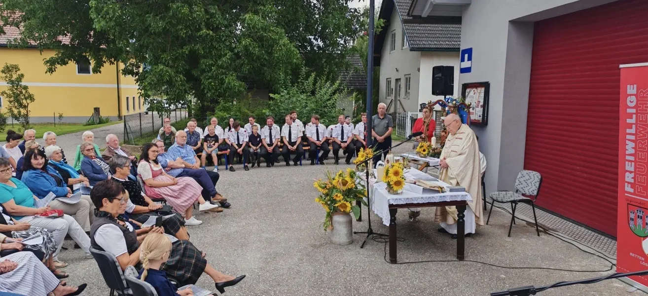 Ein Priester steht an einem Tisch mit Sonnenblumen und hält eine Rede vor einer Gruppe von uniformierten Personen, die vor einem Haus sitzen.
