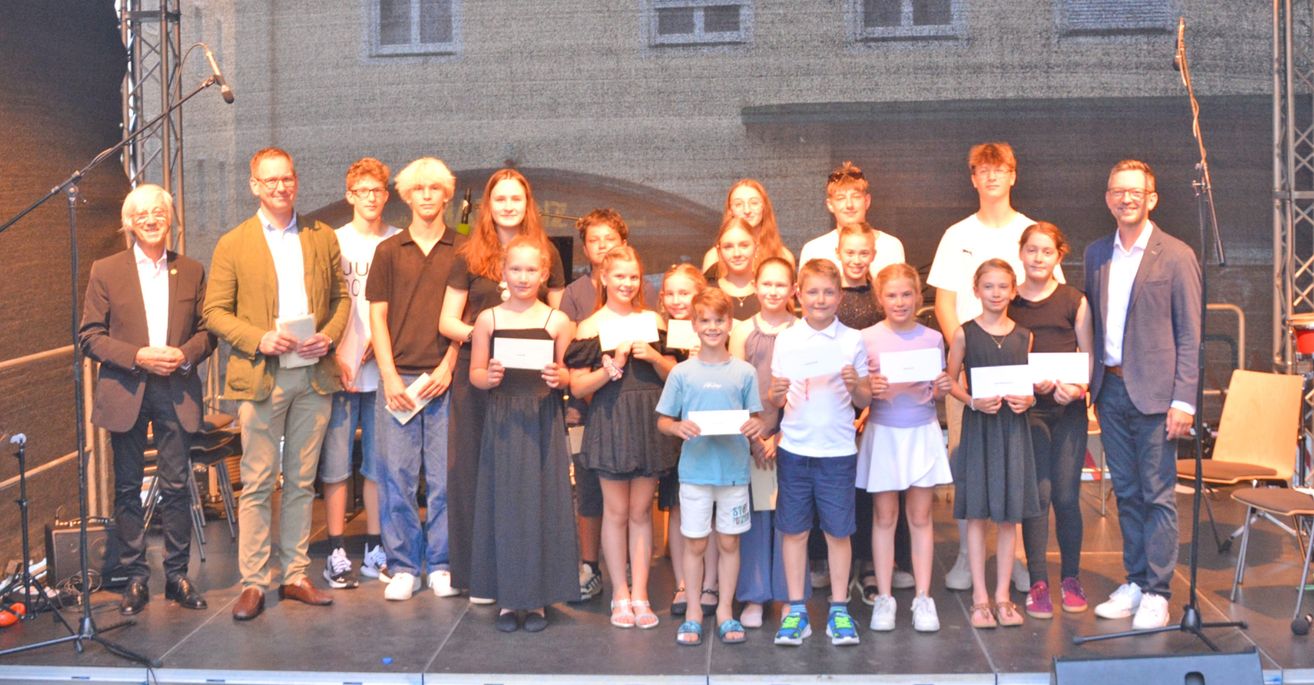 Eine Gruppe junger Menschen steht auf der Bühne und hält Urkunden in den Händen, lächelt für ein Foto. Sie sind lässig gekleidet und scheinen eine Leistung zu feiern.