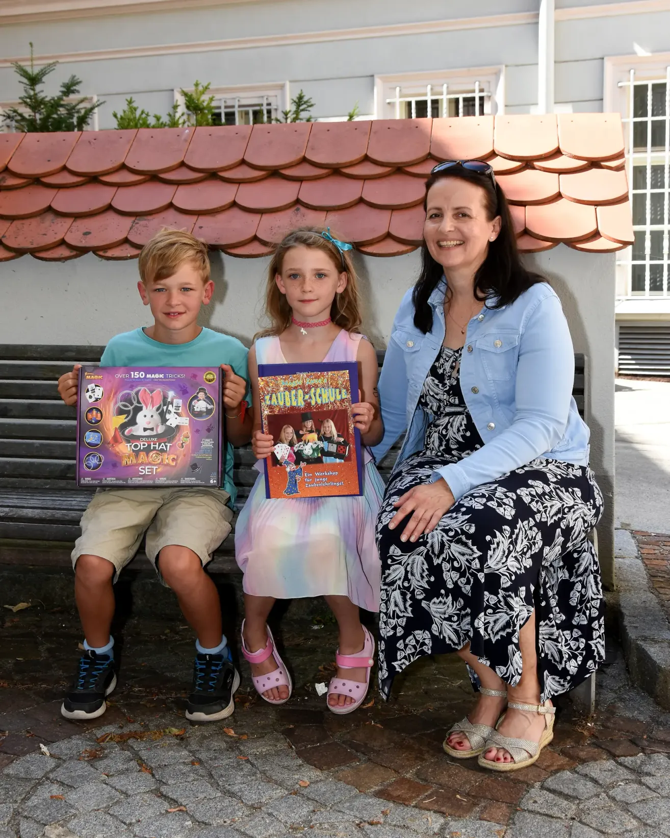 Eine Frau und zwei Kinder sitzen auf einer Bank und lächeln. Der Junge hält eine 'Top Hat Magic Set'-Box und das Mädchen eine 'Lauder-Schule'-Box. Hinter ihnen ist ein roter Dachziegel.