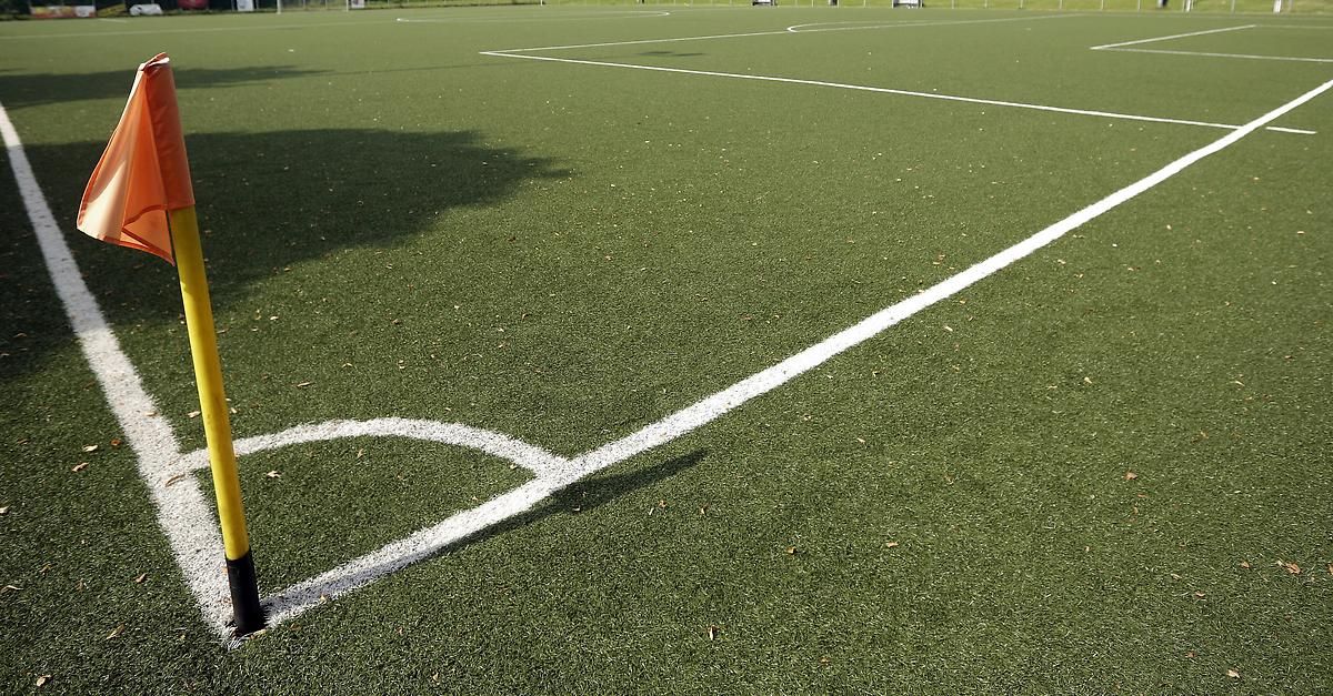 Ein leerer Fußballplatz mit weißen Linien, die die Grenzen markieren, und einem Schatten eines Baumes auf der linken Seite.