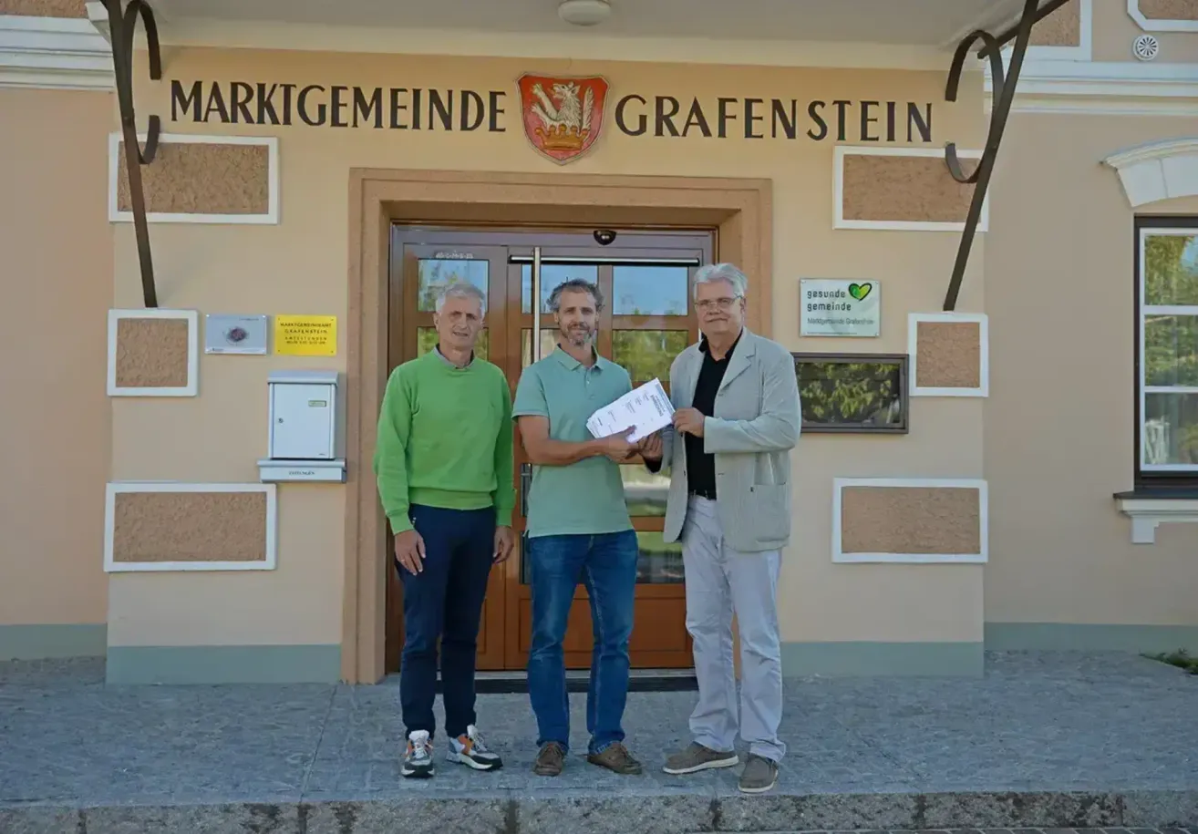 Drei Männer stehen vor einem Gebäude mit dem Namen 'Marktgemeinde Grafenstein'. Der Mann links trägt einen grünen Pullover, der in der Mitte ein grünes Poloshirt und der Mann rechts einen grauen Anzug. Sie alle lächeln und scheinen für ein Foto zu posieren.