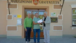 Drei Männer stehen vor einem Gebäude mit dem Namen 'Marktgemeinde Grafenstein'. Der Mann links trägt einen grünen Pullover, der in der Mitte ein grünes Poloshirt und der Mann rechts einen grauen Anzug. Sie alle lächeln und scheinen für ein Foto zu posieren.