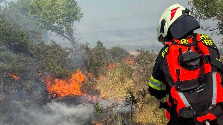 Ein Feuerwehrmann in voller Ausrüstung steht in der Nähe eines brennenden Bereichs, mit Flammen und Rauch, der aus dem Boden aufsteigt.