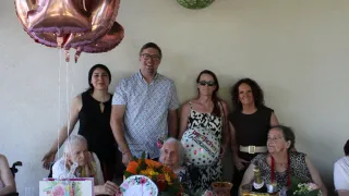 Eine Gruppe von Menschen, darunter ältere Frauen und ein Mann, versammelt sich um einen Tisch für eine Geburtstagsfeier. Sie lächeln und posieren für ein Foto mit Luftballons und einem Kuchen im Hintergrund.