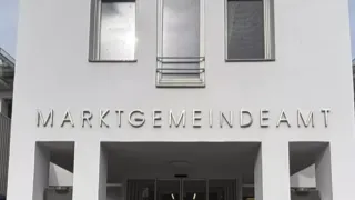Ein weißes Gebäude mit dem Namen 'ARKTGEMEINDEA' über dem Eingang, mit drei Fenstern und einer Tür mit Glasscheibe.