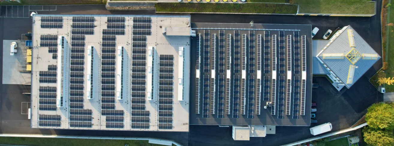 Eine Vogelperspektive eines Gebäudes mit einer großen Anordnung von Solarpanelen auf dem Dach. Die Paneele sind ordentlich in Reihen angeordnet, und das Gebäude ist von einem Parkplatz umgeben.