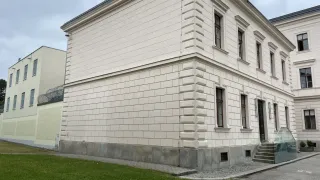 Ein großes weißes Gebäude mit mehreren Fenstern und einem Betonsockel, umgeben von einem Rasen und Stufen, die zum Eingang führen.