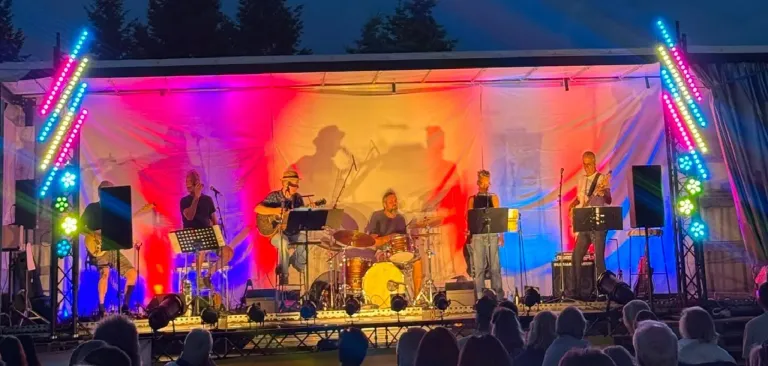 Eine Band spielt auf der Bühne unter leuchtendem Bühnenlicht, wobei ein Mitglied Gitarre spielt und ein anderes Schlagzeug. Das Publikum ist im Vordergrund sichtbar.