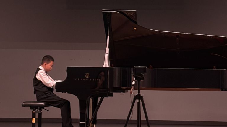 Ein junger Junge spielt ein Steinway & Sons Konzertflügel auf der Bühne, mit einem Stativ und einer Kamera in der Nähe.