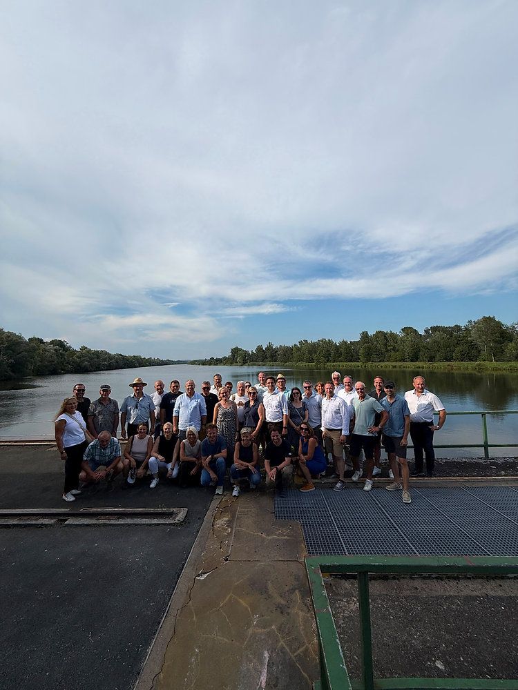 Eine Gruppe von Menschen in Freizeitkleidung posiert für ein Foto auf einer Brücke über einem Fluss mit einem bewölkten Himmel im Hintergrund.