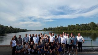 Eine Gruppe von Menschen in Freizeitkleidung posiert für ein Foto auf einer Brücke über einem Fluss mit einem bewölkten Himmel im Hintergrund.