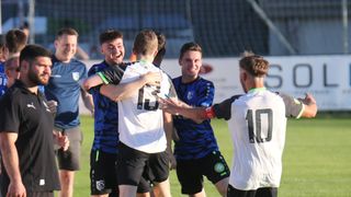Eine Gruppe junger männlicher Fußballspieler in blauen und weißen Trikots feiert auf dem Spielfeld. Sie lächeln und umarmen sich.
