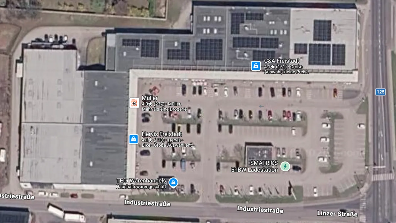 Luftaufnahme eines Industriegebiets mit mehreren Gebäuden und Parkplätzen, einschließlich eines großen Lagerhauses mit Solarpanelen. Es gibt mehrere Beschriftungen, die Unternehmen und Dienstleistungen angeben.