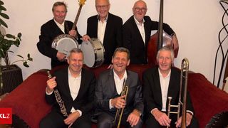Eine Gruppe von Musikern in formeller Kleidung posiert für ein Foto. Drei sitzen auf einem Sofa, und drei stehen hinter ihnen, jeder mit einem Musikinstrument.
