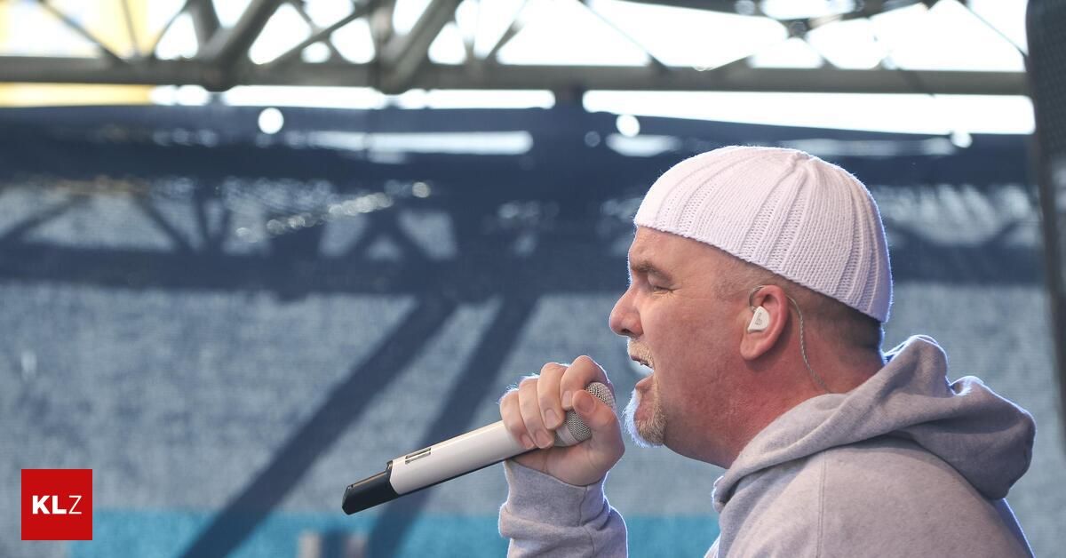 Ein Mann mit einer weißen Mütze und einem grauen Hoodie singt in ein Mikrofon auf der Bühne.