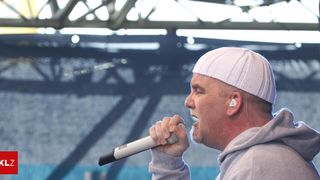 Ein Mann mit einer weißen Mütze und einem grauen Hoodie singt in ein Mikrofon auf der Bühne.