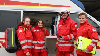Vier Personen in roten Uniformen, zwei Frauen und zwei Männer, stehen vor einem Krankenwagen, lächeln und posieren für ein Foto.